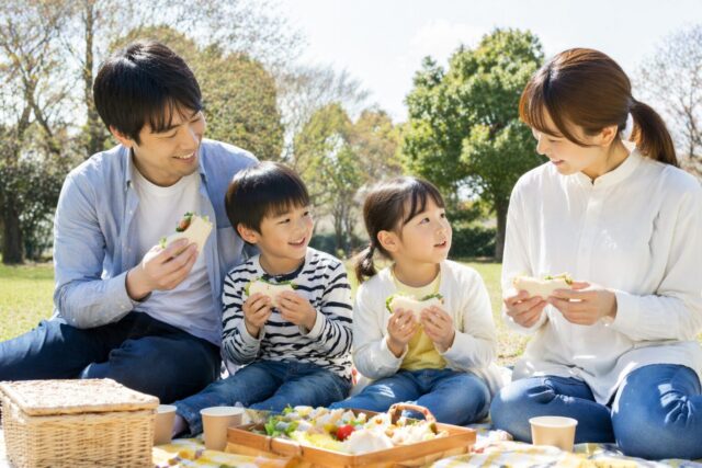 4人家族が天気の良い屋外のレジャーシートの上でのピクニックで大きな入れ物に詰められた手作りのサンドウィッチのお弁当を食べている様子